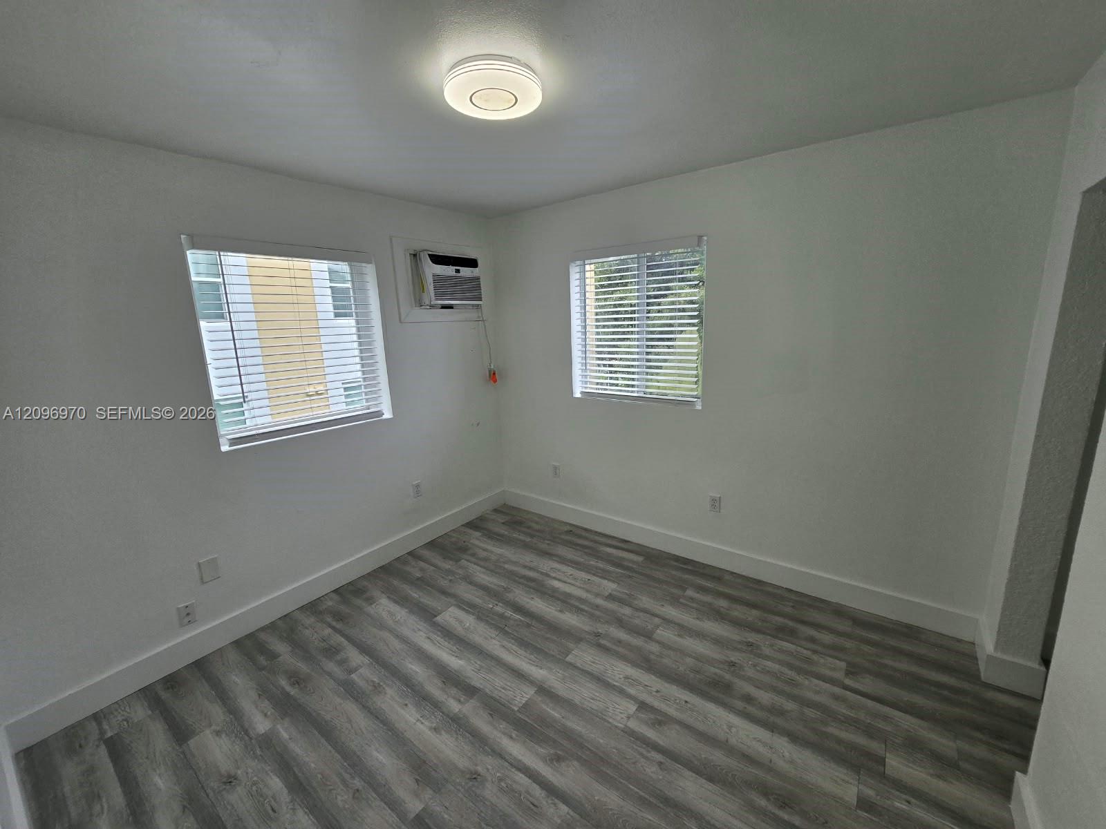 Neutral-toned wood-finish flooring, two windows with horizontal blinds, flush-mount ceiling light fixture, and wall-mounted air conditioning unit