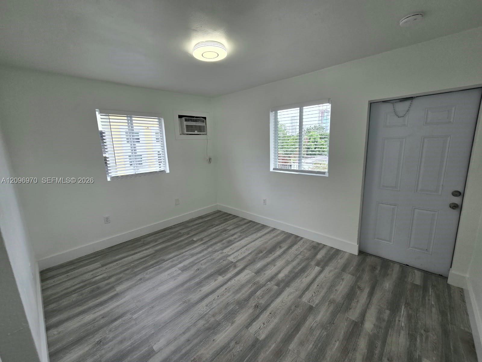 Interior space featuring wood-finish flooring, white walls, dual windows with blinds, a flush-mount ceiling light fixture, and a six-panel entry door Interior space featuring wood-finish flooring, white walls, dual windows with blinds, a flush-mount ceiling light fixture, and a six-panel entry door