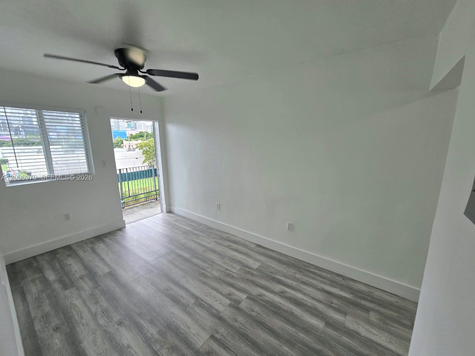 Interior space featuring wood-finish flooring, a multi-blade ceiling fan with integrated light fixture, a window with horizontal blinds, and access to a balcony with metal railing Interior space featuring wood-finish flooring, a multi-blade ceiling fan with integrated light fixture, a window with horizontal blinds, and access to a balcony with metal railing