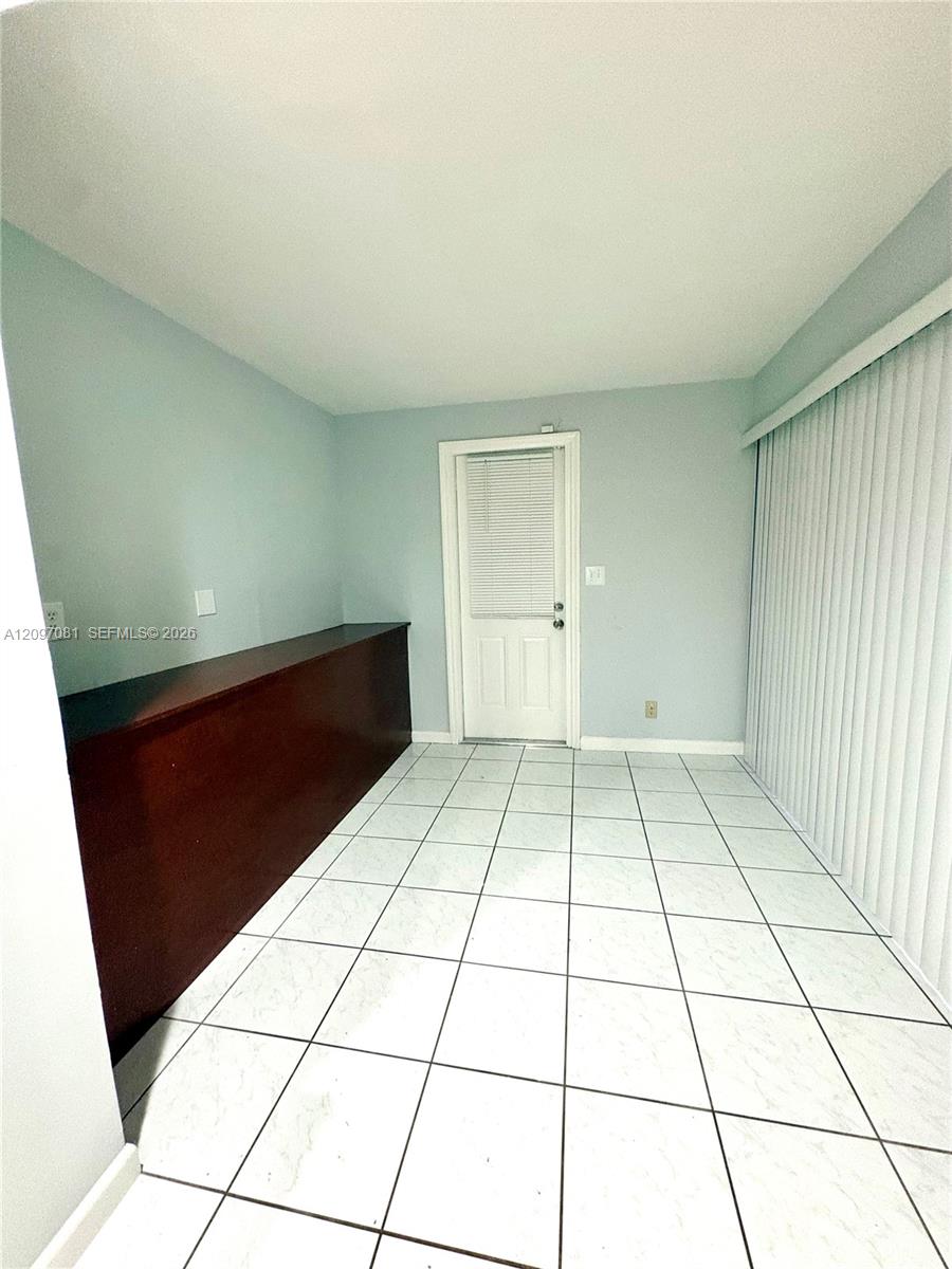 Room featuring light-colored tile flooring, built-in custom cabinetry with a wood-tone finish, a single-panel exterior door, and large vertical blinds