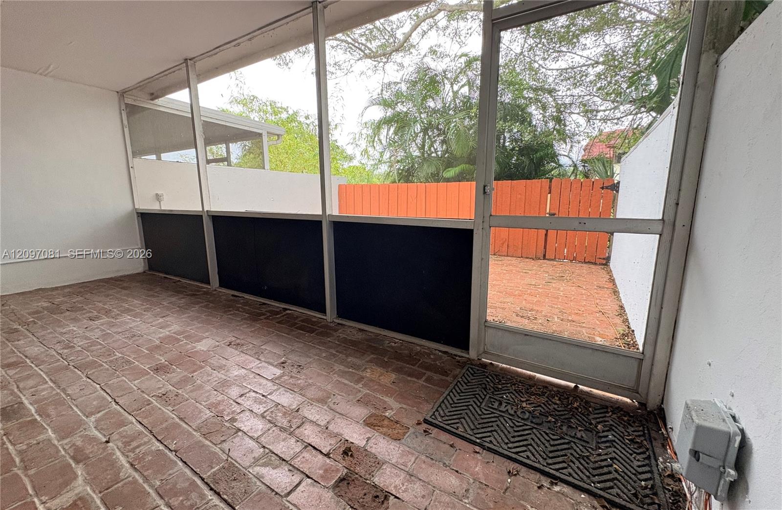 Screened-in patio featuring brick flooring, white wall finishes, and a swinging aluminum-frame door opening to a private outdoor space with a privacy fence