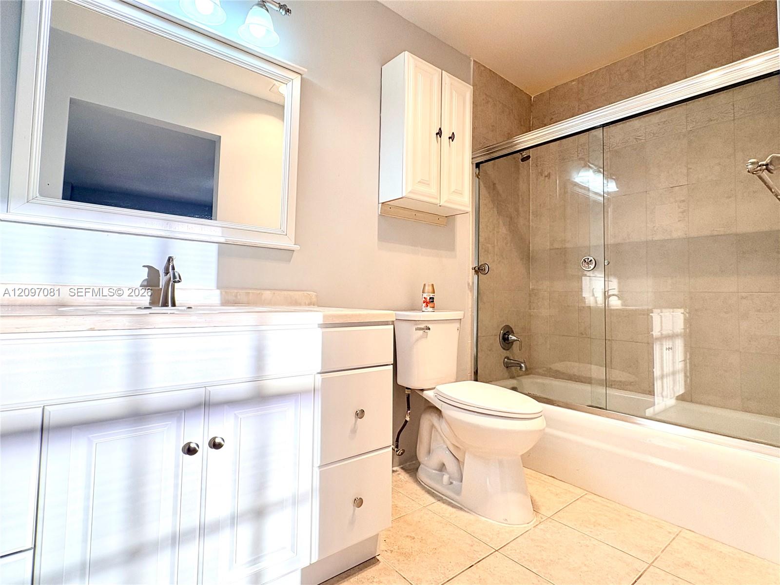 Bathroom featuring white shaker-style cabinetry, framed vanity mirror, wall-mounted storage cabinet, tile-surround tub with sliding glass doors, and light-toned floor tile