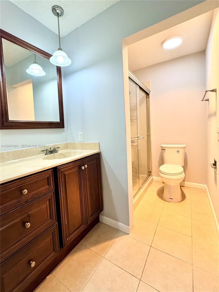 Bathroom vanity featuring wood cabinetry, stone-finish countertop, undermount sink, and wall-mounted pendant lighting, accompanied by a walk-in shower with a glass enclosure and a ceramic tile floor