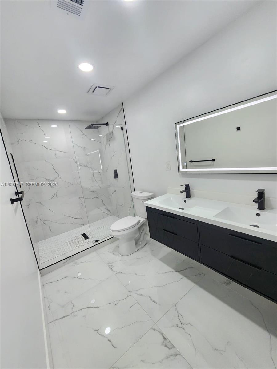 Floating double vanity with matte black faucets, oversized walk-in shower with glass enclosure, wall-to-wall stone-finish tile flooring, and integrated LED-backlit mirror