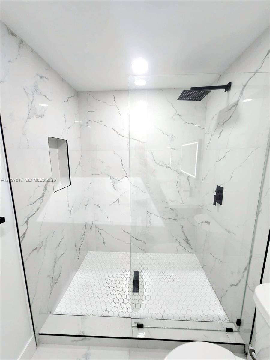 Walk-in shower featuring large-format white stone-finish wall tile, hexagonal floor tiling, a frameless glass enclosure, a built-in recessed niche, and matte black plumbing fixtures
