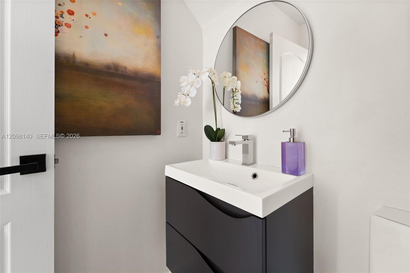 Modern wall-mounted dark cabinetry with a white integrated sink, polished chrome single-handle faucet, and circular frameless mirror
