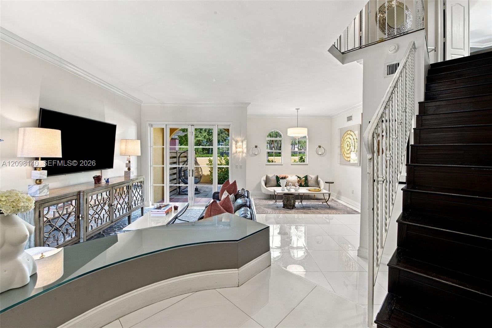 Open-concept living space featuring high-gloss tile flooring, an architectural curved built-in desk with glass surface, a dark-stained wood staircase with decorative metal railing, and multi-pane glass French doors