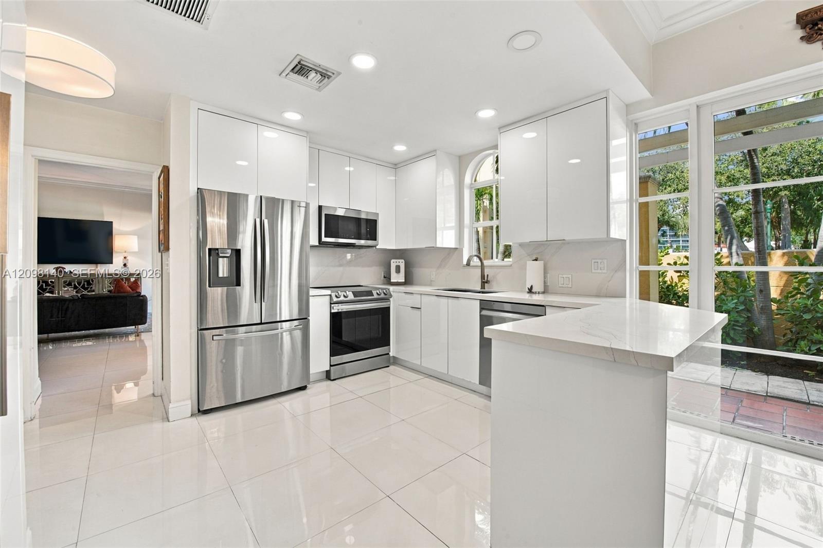 Kitchen featuring high-gloss white cabinetry, stone-finish countertops, stainless steel appliances, large-format tile flooring, and floor-to-ceiling glass paneling
