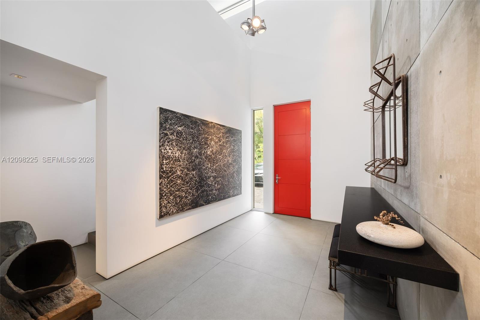High-ceiling entryway featuring large-format tile flooring, a prominent red entry door, a floor-to-ceiling vertical window, and a feature wall composed of modular concrete panels with a built-in black console shelf