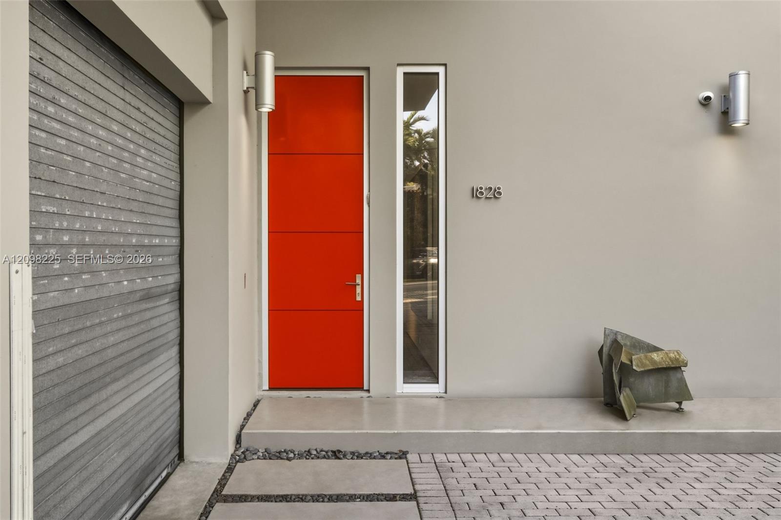 Modern exterior entry featuring a high-contrast orange door, tall side-light window, metallic wall-mounted light fixtures, and a paved brick driveway