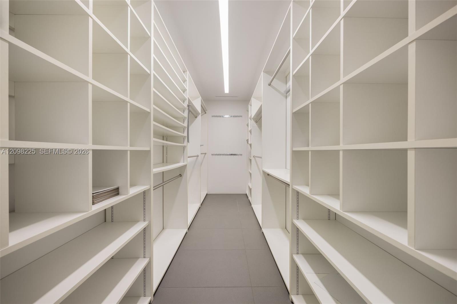 Custom walk-in storage featuring floor-to-ceiling white shelving units, integrated hanging rods, and large-format tile flooring with linear overhead lighting