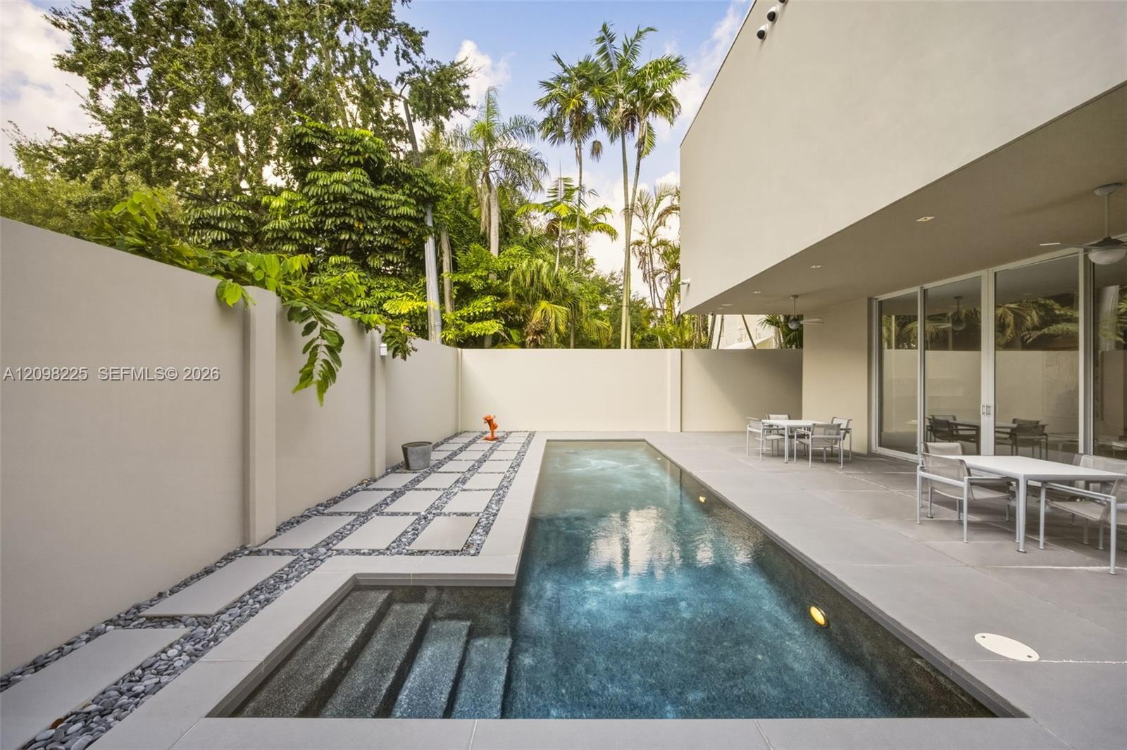 Rectangular swimming pool featuring integrated steps, surrounded by large-format concrete patio tiling, decorative stone-accented pathways, and solid perimeter wall fencing