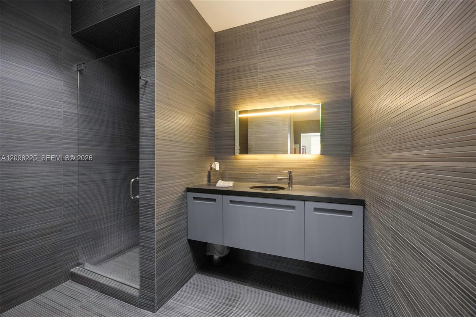 Modern bathroom featuring floor-to-ceiling linear-textured wall tile, a floating vanity with a solid surface countertop, a built-in backlit mirror, and a glass-enclosed shower stall