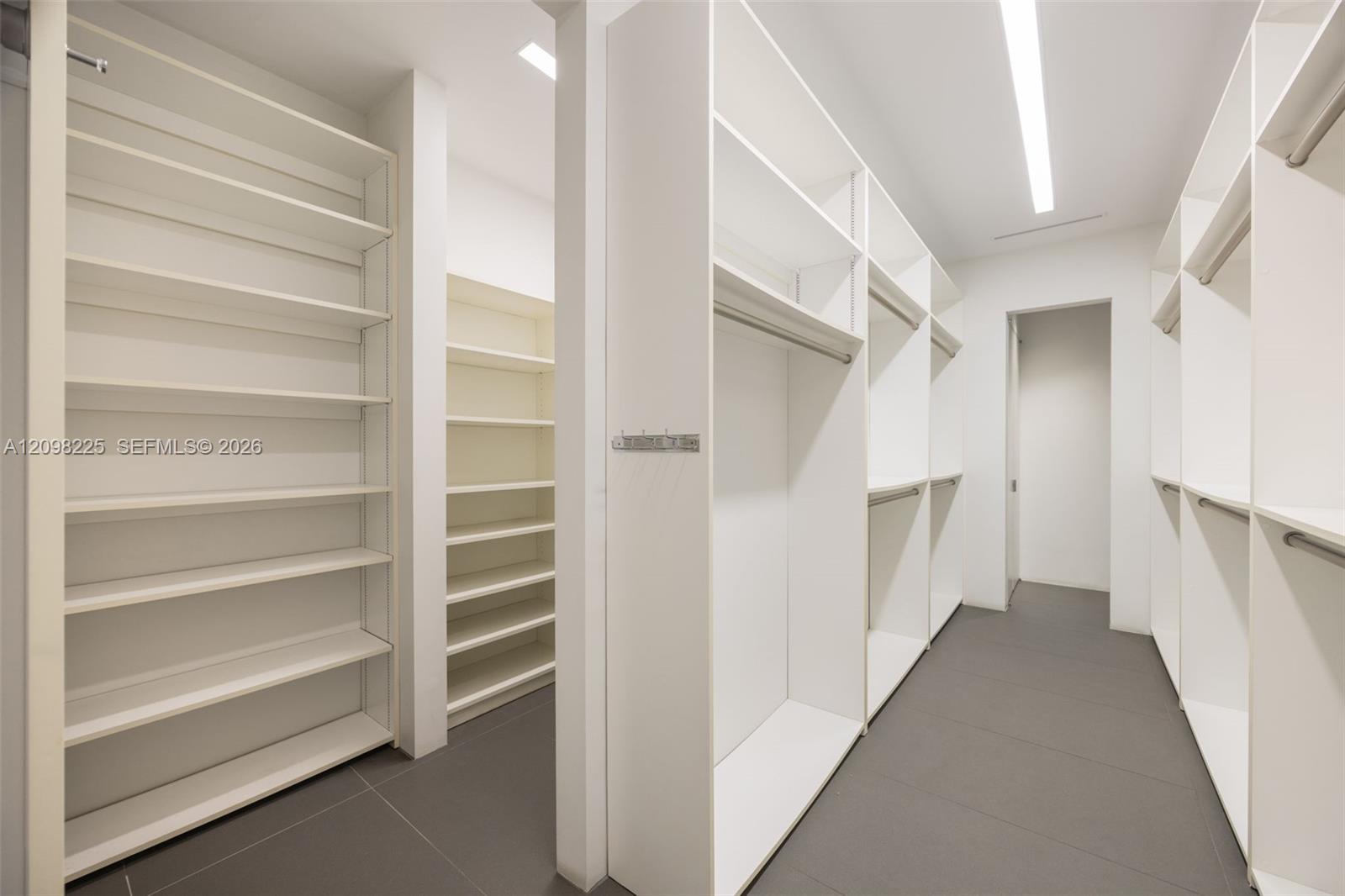 Custom walk-in closet featuring floor-to-ceiling white shelving, integrated hanging rods, minimalist cabinetry, and large-format tile flooring with recessed linear lighting