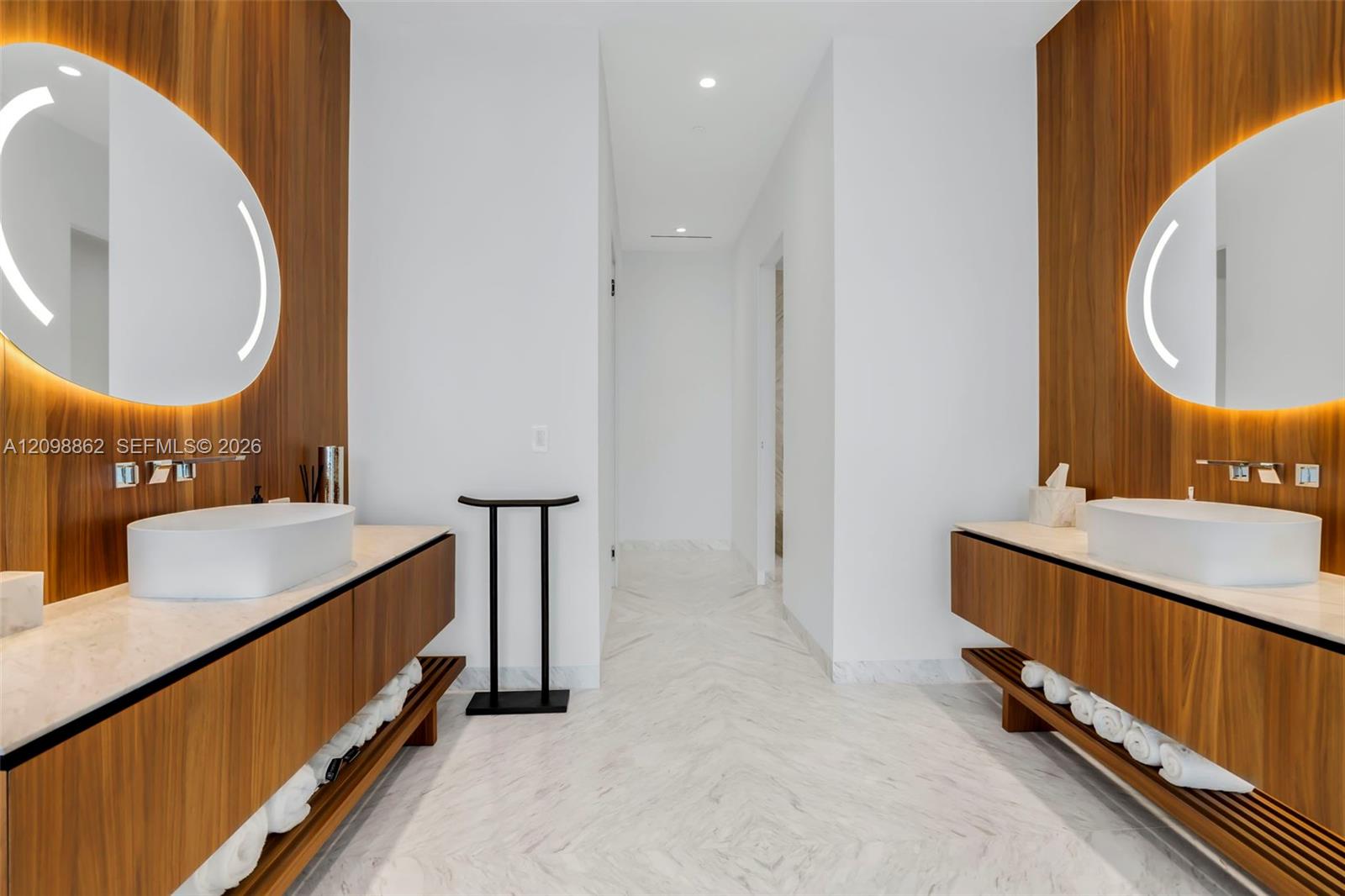 Dual floating wood-finish vanities with stone countertops, integrated basin sinks, wall-mounted faucets, oval backlit mirrors, and stone flooring in a herringbone pattern