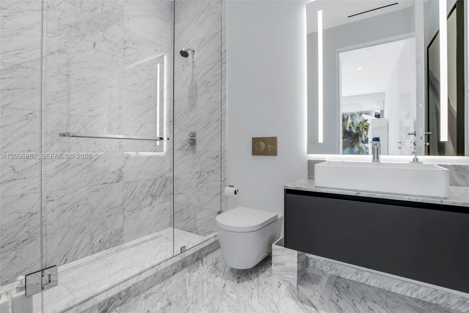 Full-height stone wall tiling, glass-enclosed walk-in shower, wall-mounted commode with metallic flush plate, floating vanity with rectangular vessel sink, and integrated LED-backlit mirror