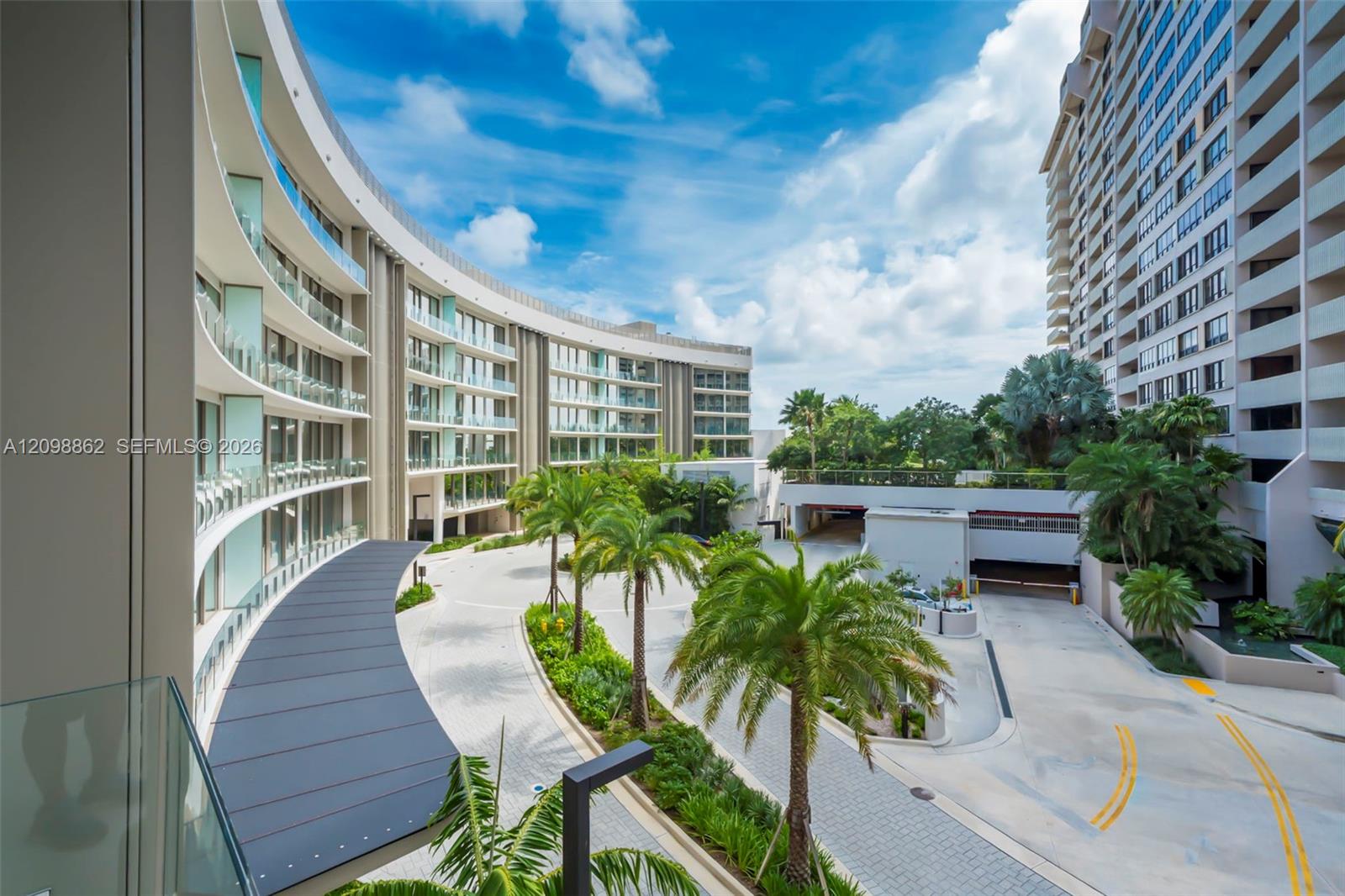 Curved multi-story residential architecture featuring glass balcony railings, covered arrival canopy, paved motor court, and mature palm landscaping