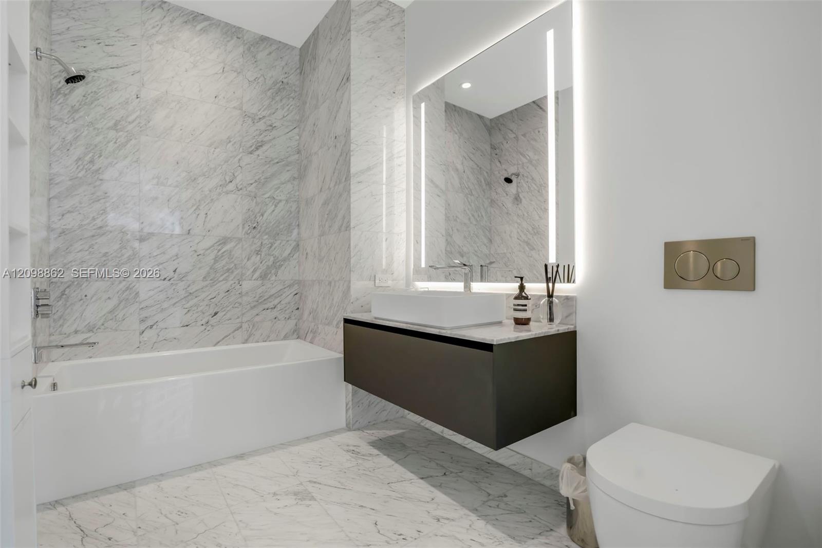 Full-height natural stone tiling in shower and tub surround, floating vanity with rectangular vessel sink, wall-mounted faucet, integrated LED-lit vanity mirror, and wall-mounted dual-flush toilet