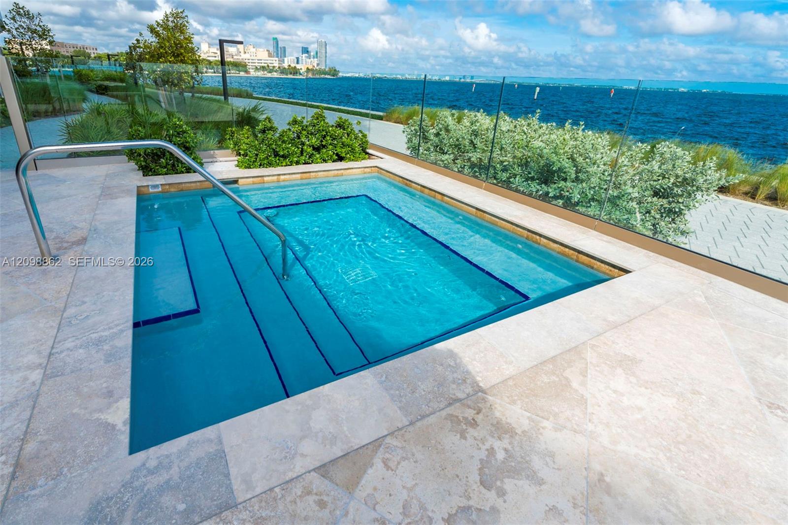 In-ground spa with integrated staircase and stainless steel handrails, surrounded by stone paver decking and a glass perimeter railing, overlooking a coastal waterfront