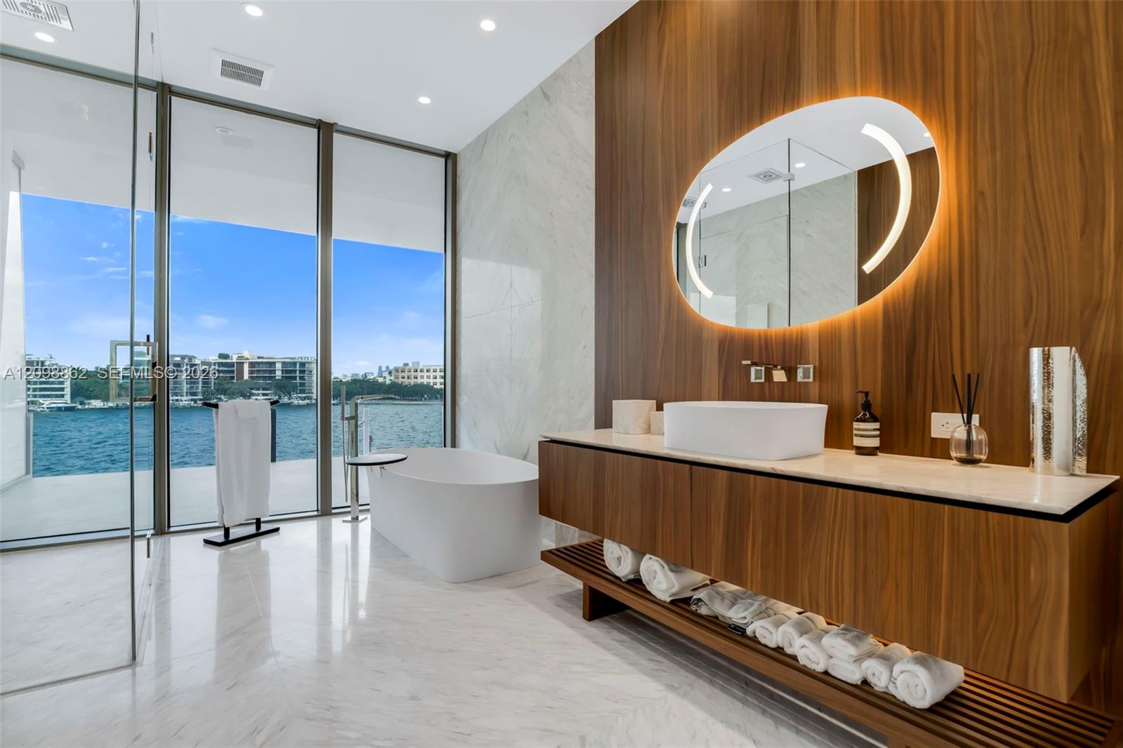 Floating wood-finish vanity with countertop basin, integrated LED-backlit mirror, floor-to-ceiling glass paneling, and freestanding soaking tub