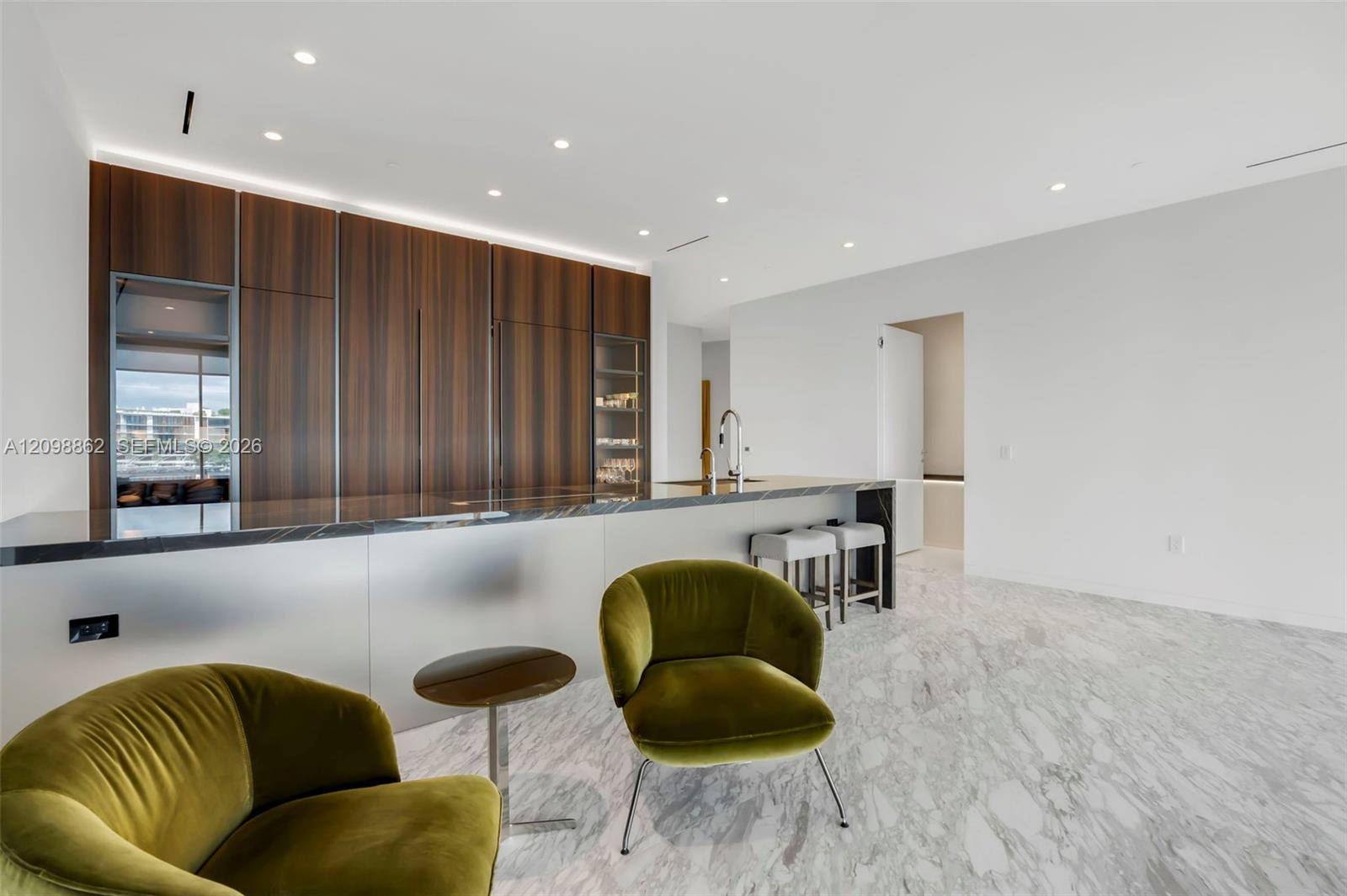 Modern kitchen featuring floor-to-ceiling wood-finish cabinetry, a large stone-surfaced island with integrated sink, recessed ceiling lighting, and light-toned stone flooring