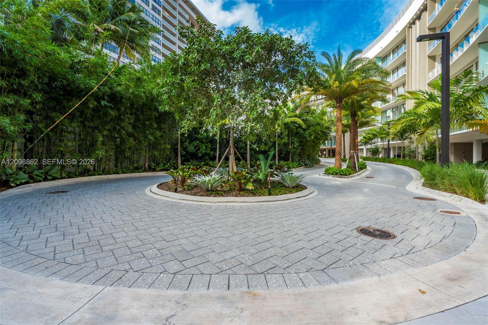 Circular motor court featuring stone paver driveway, central island landscaping, mature palm trees, and modern exterior architecture