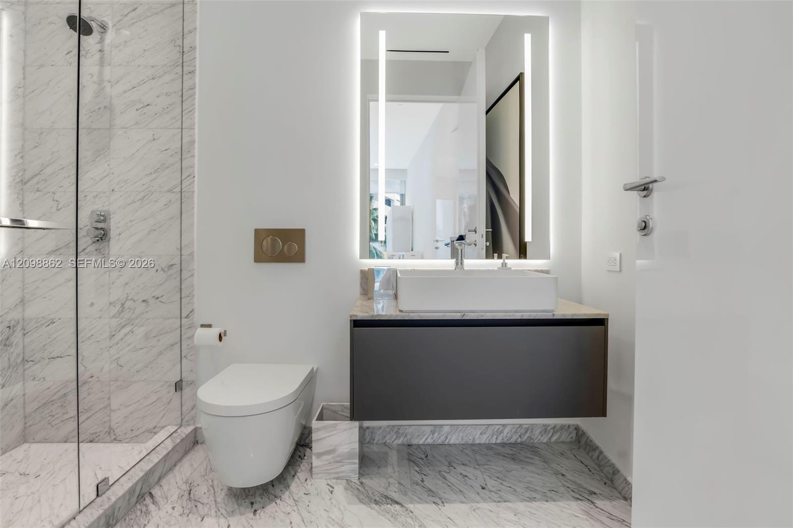 Bathroom featuring a glass-enclosed shower with stone tile surround, floating vanity with a rectangular vessel sink, wall-mounted toilet, illuminated mirror, and stone flooring