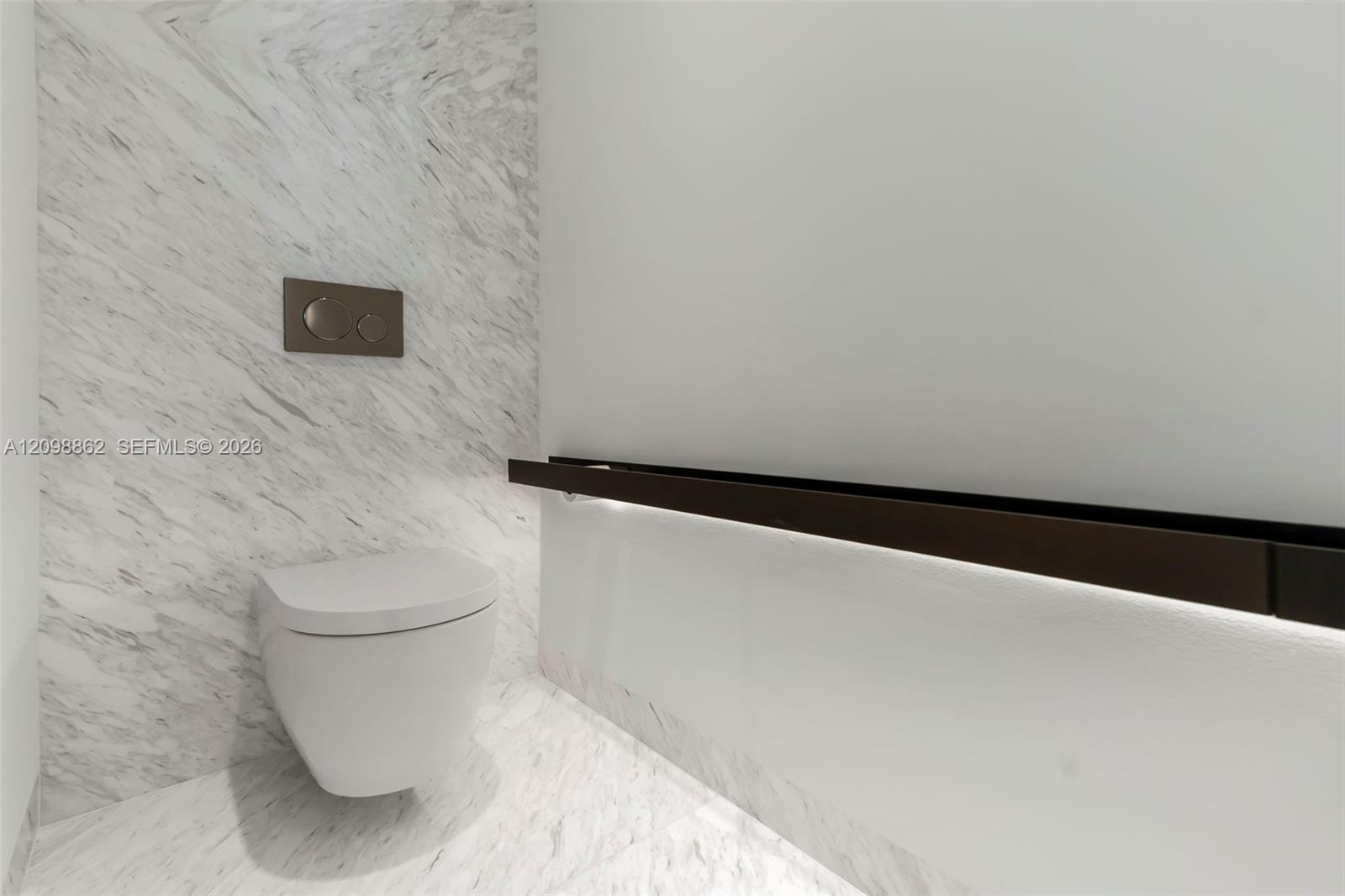 Wall-mounted toilet with stone-finish wall cladding and floor tile, featuring a dual-flush button and a horizontal dark metal accent rail