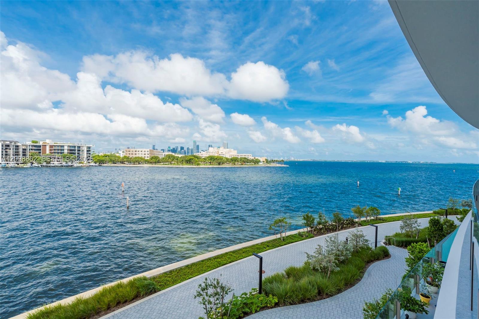 Exterior balcony featuring glass railing, paved pedestrian walkway, manicured landscaping, and direct water frontage