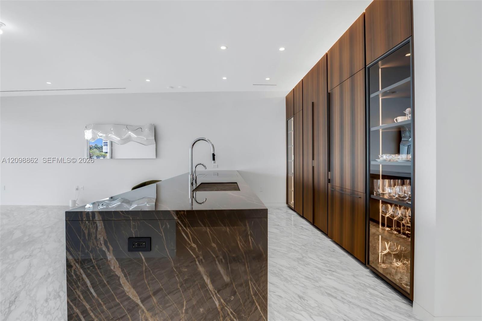 Kitchen featuring a stone-topped island with an integrated sink, full-height wood-finish cabinetry, recessed lighting, and a glass-front display cabinet Kitchen featuring a stone-topped island with an integrated sink, full-height wood-finish cabinetry, recessed lighting, and a glass-front display cabinet