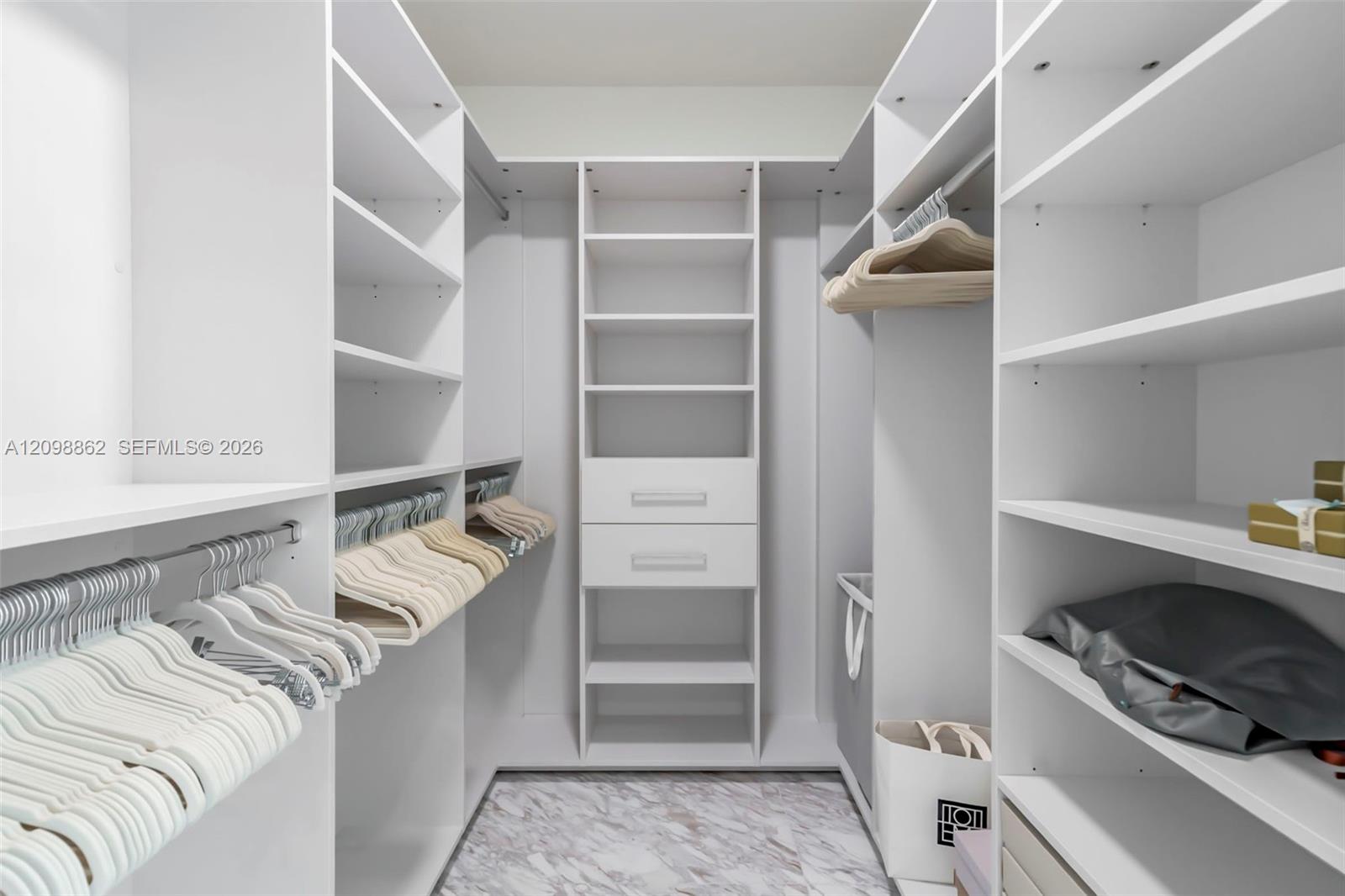 Walk-in closet featuring white built-in shelving, integrated storage drawers with hardware, overhead hanging rods, and stone-pattern flooring