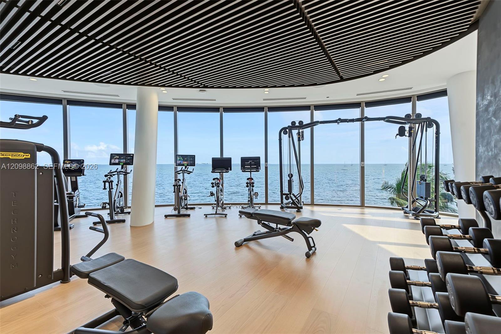 Floor-to-ceiling glass walls, expansive curved room layout, suspended linear slat ceiling, light-toned wood-finish flooring, and structural column supports