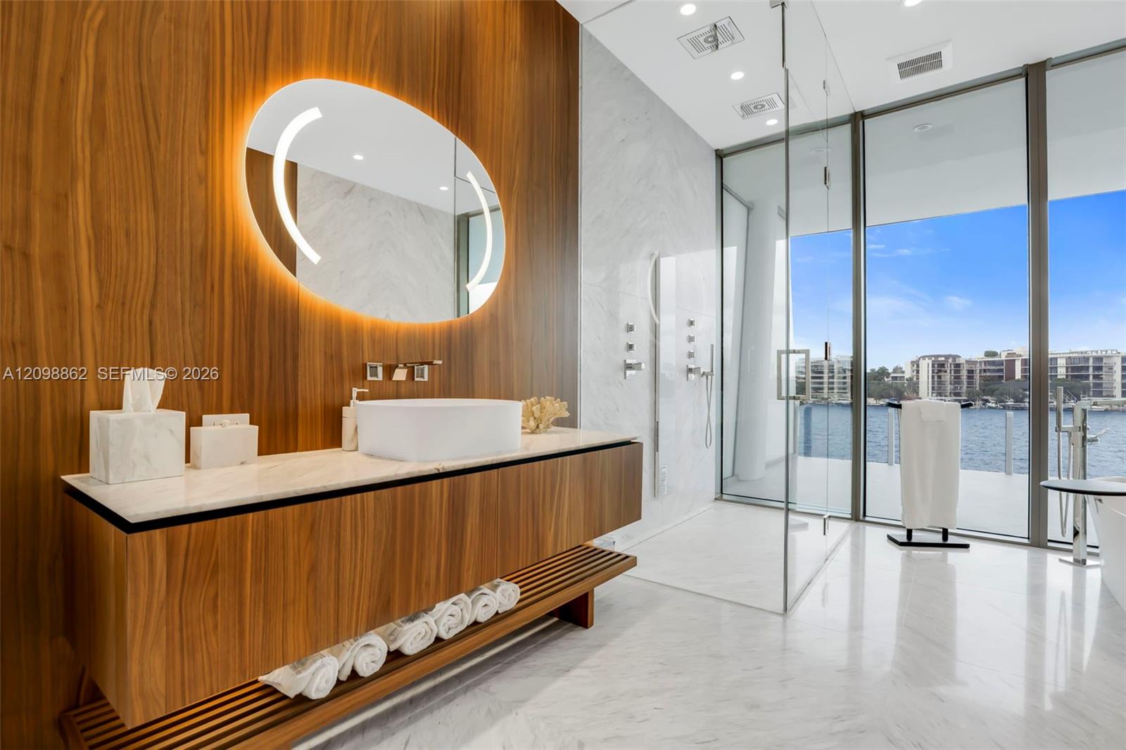 Custom wood-finish vanity with a stone countertop and vessel sink, illuminated oval mirror, stone-finish flooring, glass-enclosed shower, and full-height windows