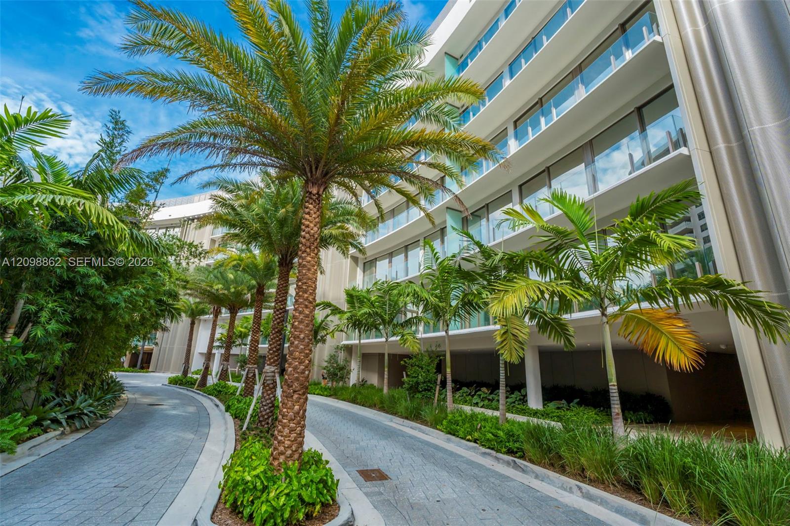 Modern multi-story facade featuring glass railings and floor-to-ceiling windows, complemented by a curved paver motor court and lush tropical landscaping