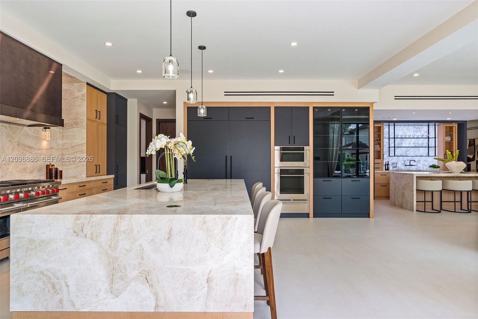Kitchen featuring a waterfall-edge island with stone surfacing, stainless steel double wall ovens, paneled cabinetry, glass-front display shelving, and pendant lighting