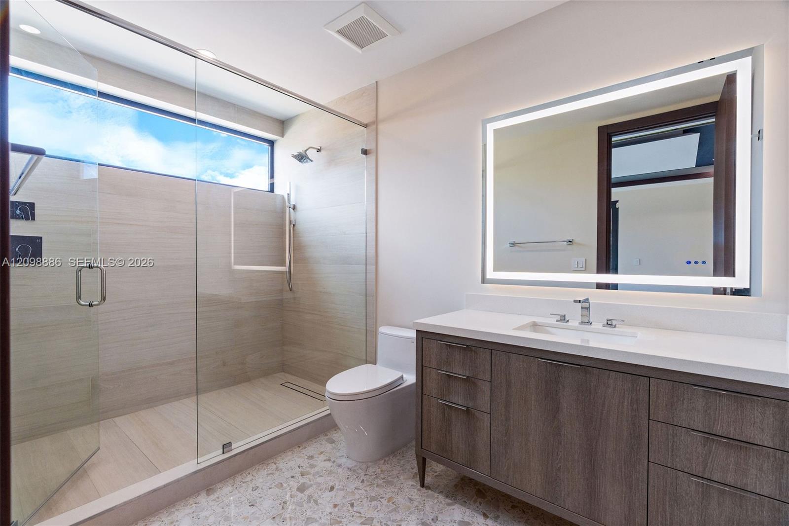 Bathroom featuring a walk-in shower with large-format tile surround, frameless glass enclosure, a wood-finish vanity with a stone countertop, and an integrated LED-lit vanity mirror