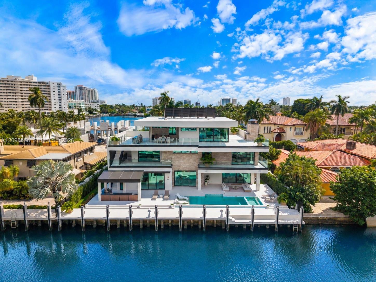 Modern waterfront residence featuring clean-lined architecture with multi-level design, expansive glass walls and sliding doors, open-air balconies with metal railings, direct canal frontage with private dock, and mature landscaping including palm trees and established greenery throughout the grounds