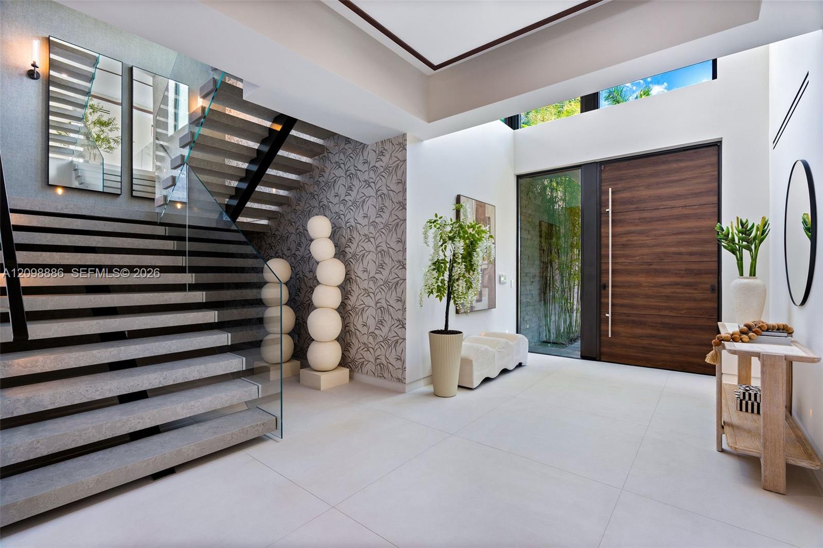 Grand foyer featuring large-format tile flooring, a custom wood-finished entry door with a vertical pull handle, a dramatic staircase with glass railings, and a double-height ceiling with clerestory windows