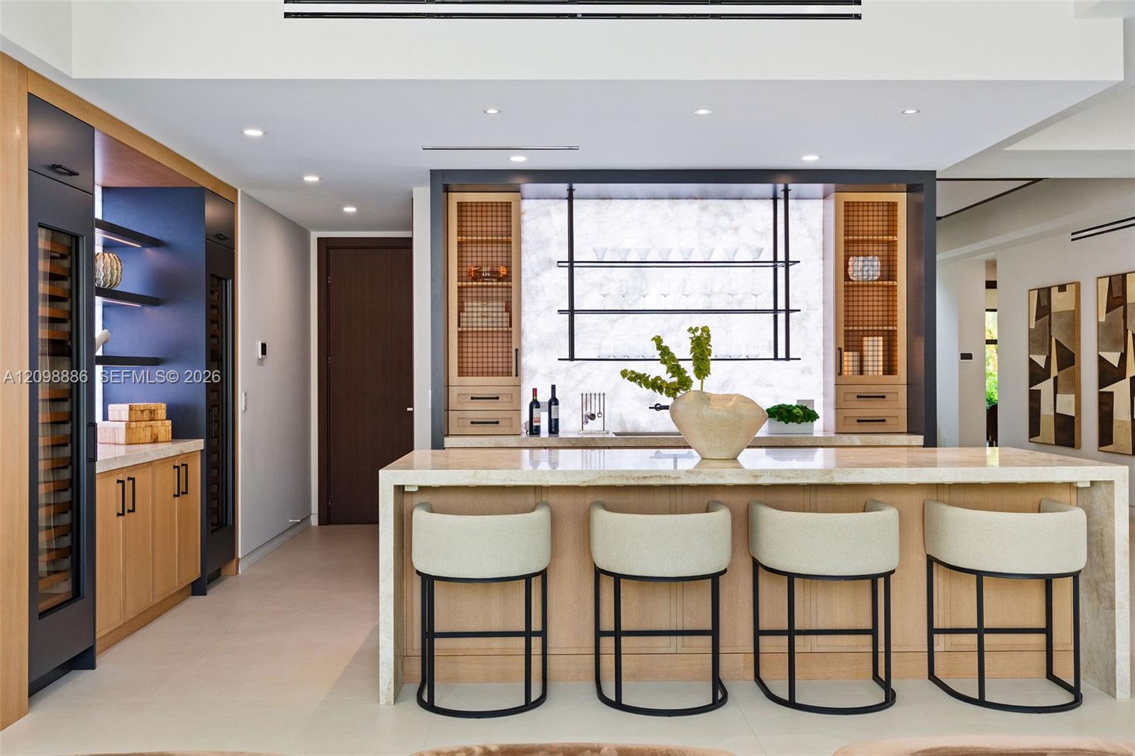 Custom bar area featuring stone slab backsplash, integrated wine refrigeration, built-in light wood cabinetry, stone-topped island with waterfall edge, and tiered floating shelving
