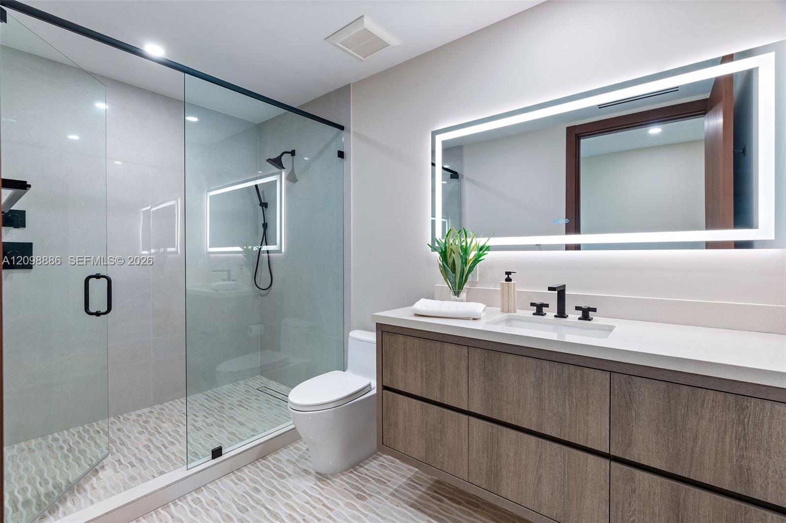 Modern bathroom featuring a wood-finish floating vanity with a white countertop, walk-in glass-enclosed shower with matte black fixtures, and a large backlit wall-mounted mirror