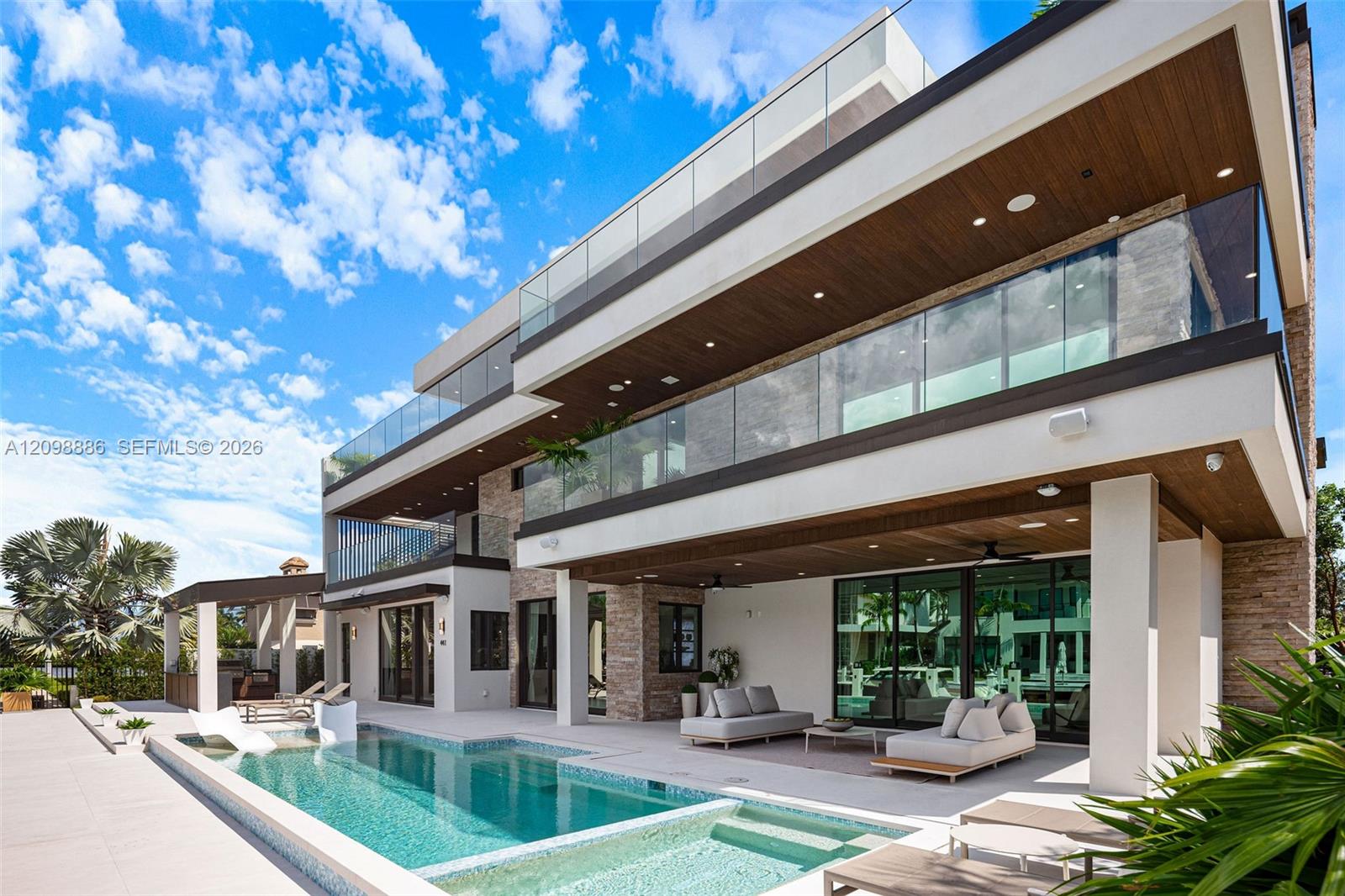 Contemporary multi-level exterior featuring stacked stone masonry, a glass-railed balcony, deep recessed wood-finish soffits, and a rectangular swimming pool with an integrated tanning ledge