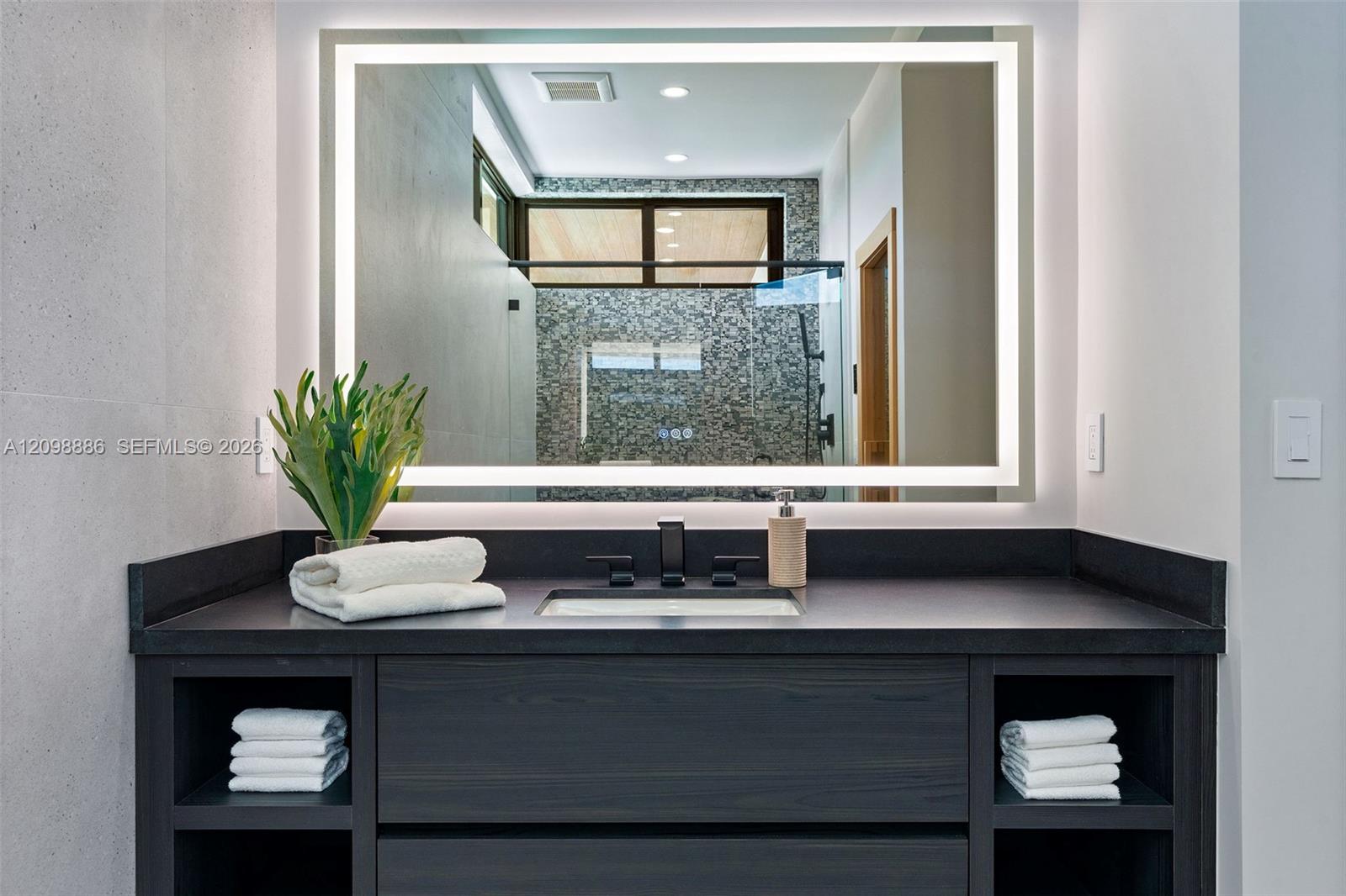 Dark wood vanity featuring integrated open shelving, a dark solid-surface countertop, an undermount rectangular sink, and a matte black faucet paired with a rectangular LED-backlit mirror