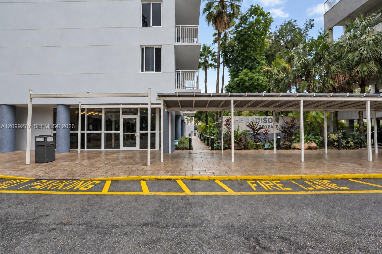 Welcoming building entrance with covered drop-off access, glass entry doors, and lush tropical landscaping surrounding The Madison.