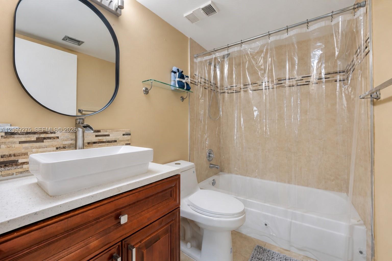 Spacious bathroom featuring a white bathtub with tiled surround, modern vessel sink, rich wood vanity, round mirror, and glass display shelves.