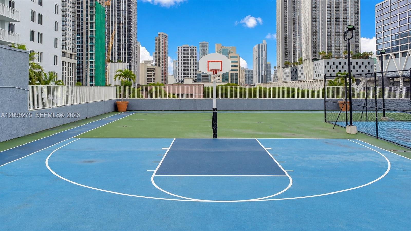 Outdoor basketball court with a blue playing surface, surrounded by modern high-rise residences and rooftop tennis facilities, offering residents convenient access to recreational amenities and downtown Miami skyline views.