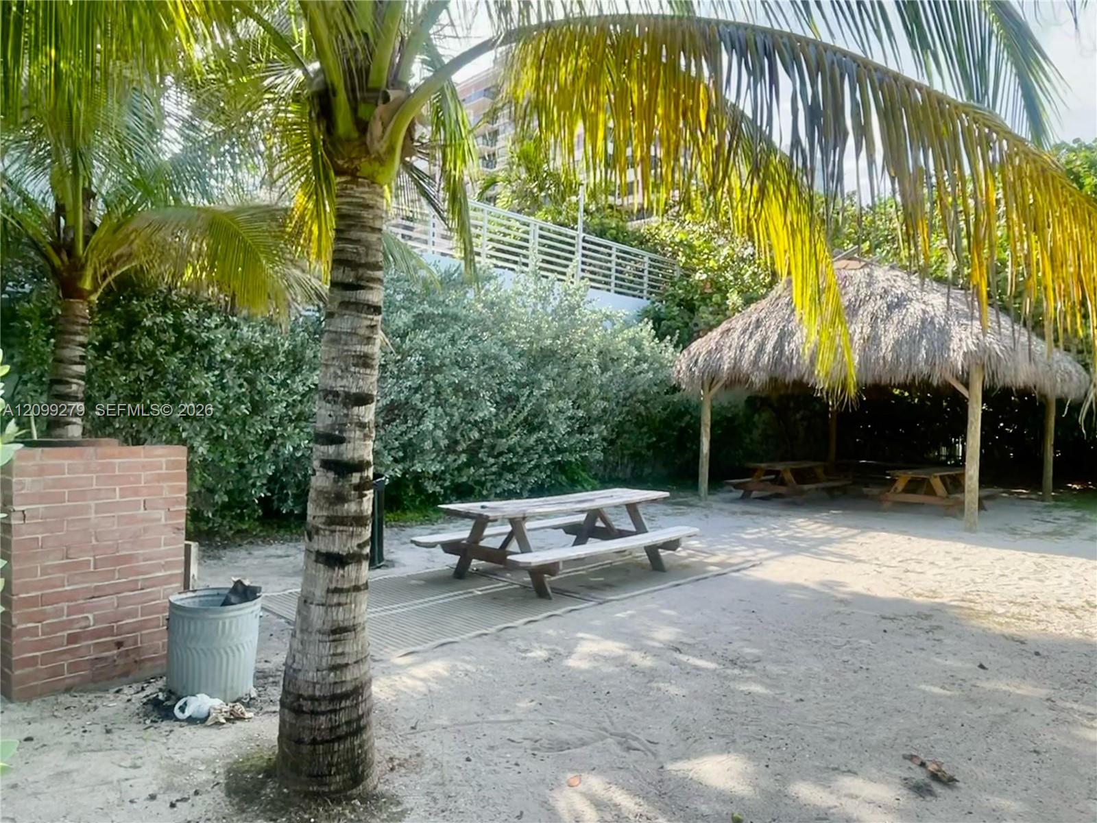 Outdoor common area featuring a thatched-roof tiki hut, sandy grounds, brick masonry structure, and mature palm trees