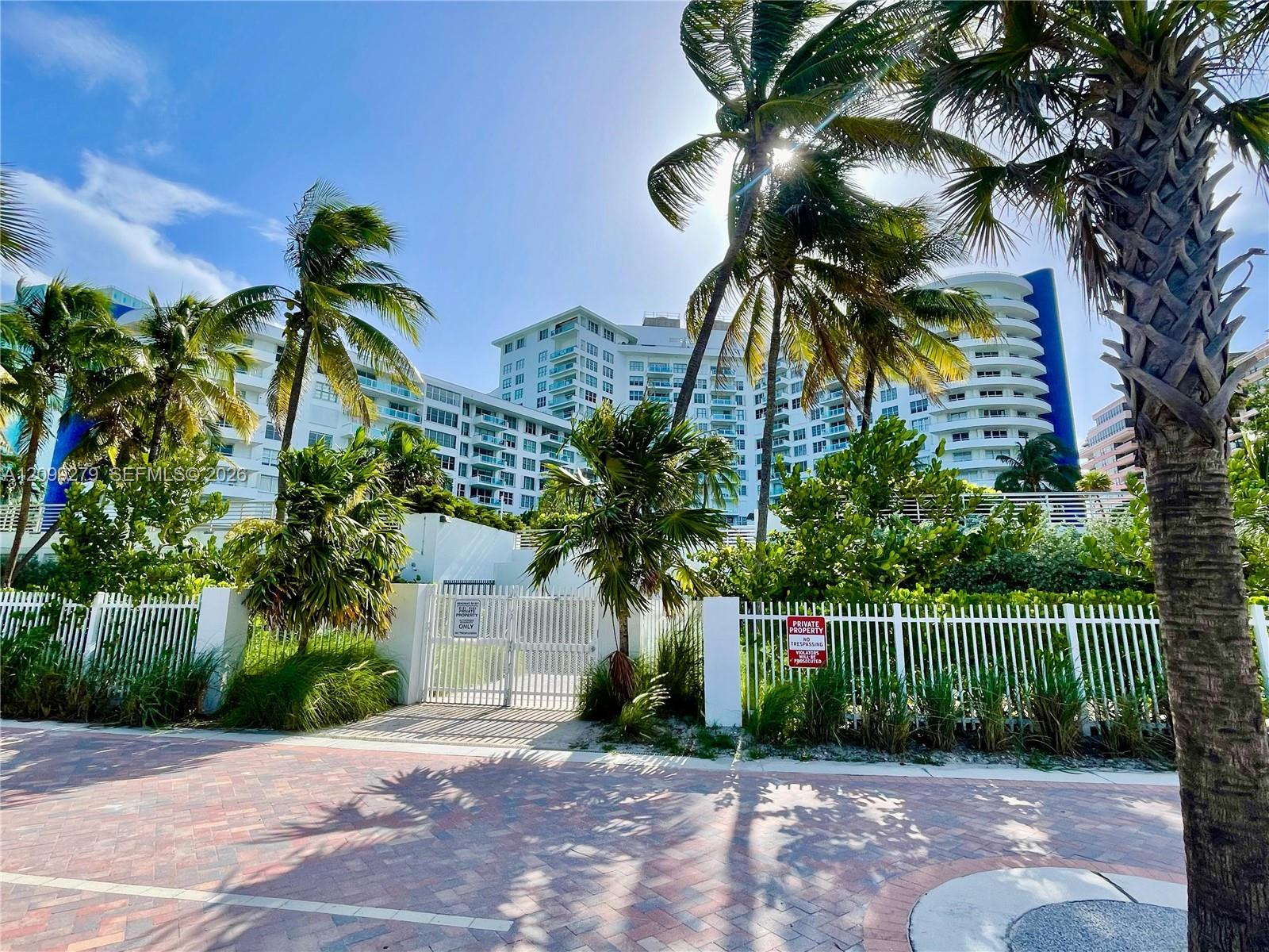 High-rise building with rounded architectural detailing, white metal security fencing, paved brick pedestrian path, and mature palm trees