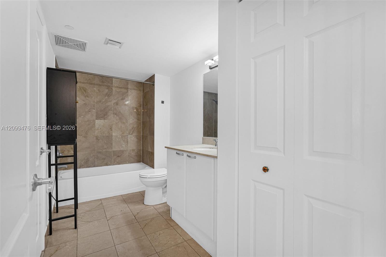 Bathroom featuring a white vanity cabinet with a solid-surface countertop, tiled tub surround, neutral-toned floor tile, and a multi-panel door closet