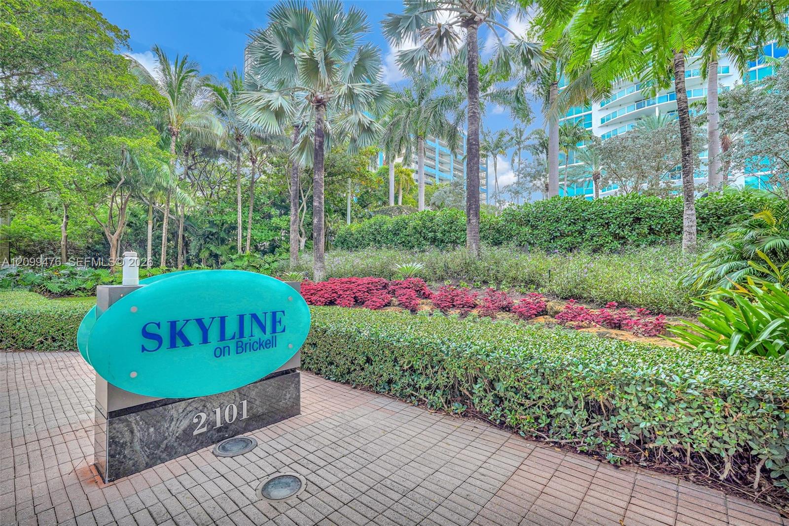 Custom property monument sign featuring a stone base and tropical landscaping including mature palm trees, tiered hedges, and perennial plantings set against a paved brick pathway