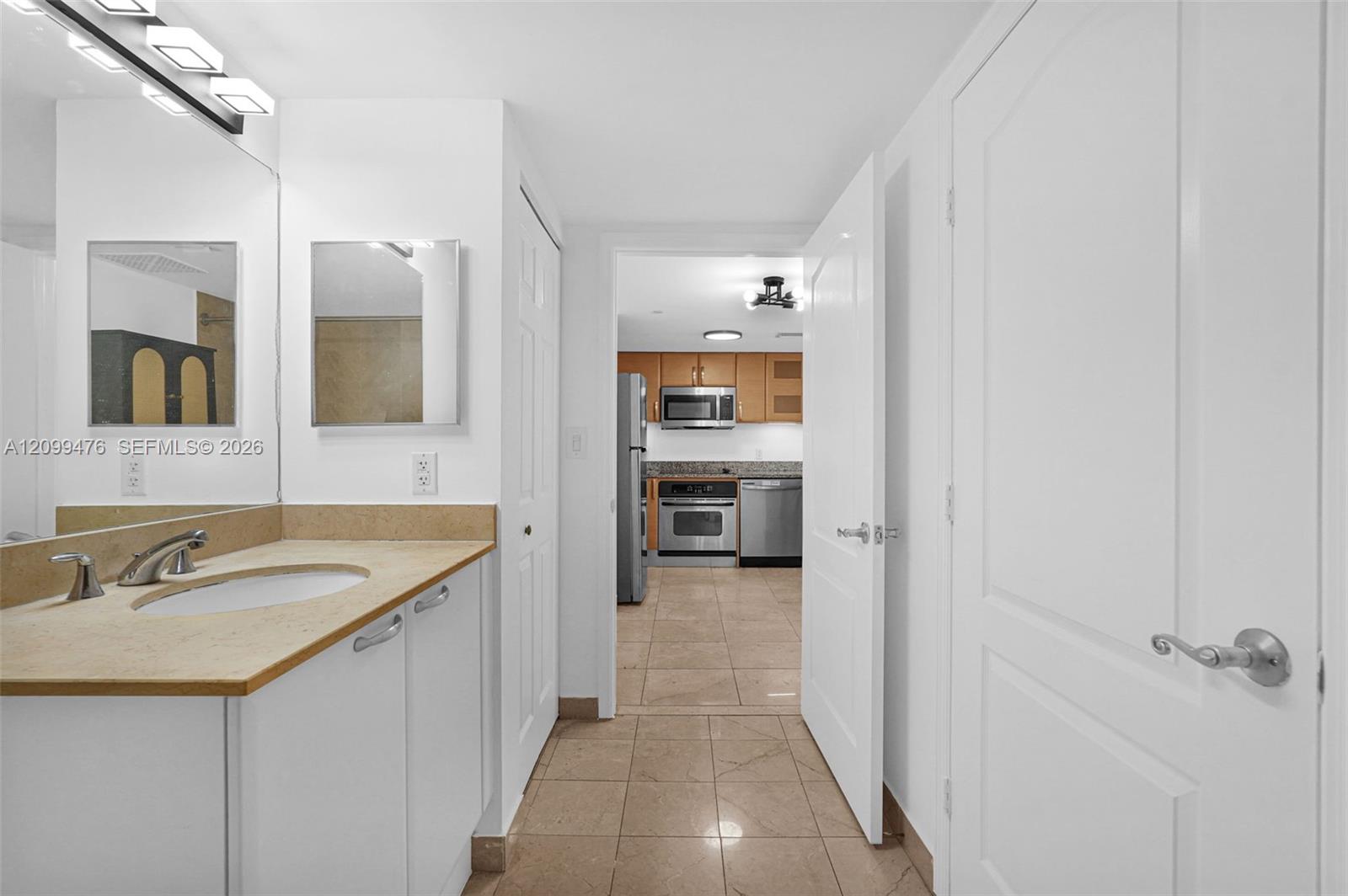 Bathroom vanity with a stone countertop, an integrated sink, a full-width mirror, and tile flooring, including an open doorway to a kitchen area featuring stainless steel appliances and wood-finish cabinetry